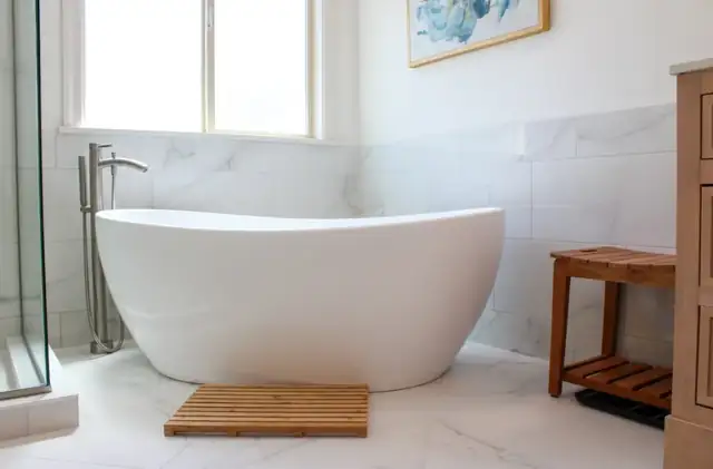 Freestanding soaking tub with floor-mounted faucet beside marble-look wall tile and a teak bath stool