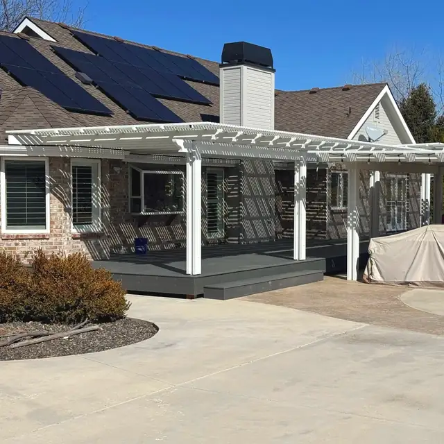 White pergola-style patio cover over a gray deck on a brick ranch home with solar panels