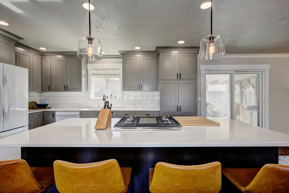 Kitchen remodeling Caldwell area: gray shaker cabinets, navy island with white quartz top, gas cooktop, glass pendants and mustard leather stools